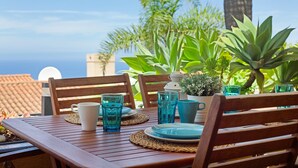 A wooden table set with plates, glasses, and a white mug, surrounded by chairs, with a view of the sea and greenery in the background. - Vacation Apartment 'Casa Ana' with Sea and Teide View, Shared Pool and Wi-Fi (SANTA URSULA)