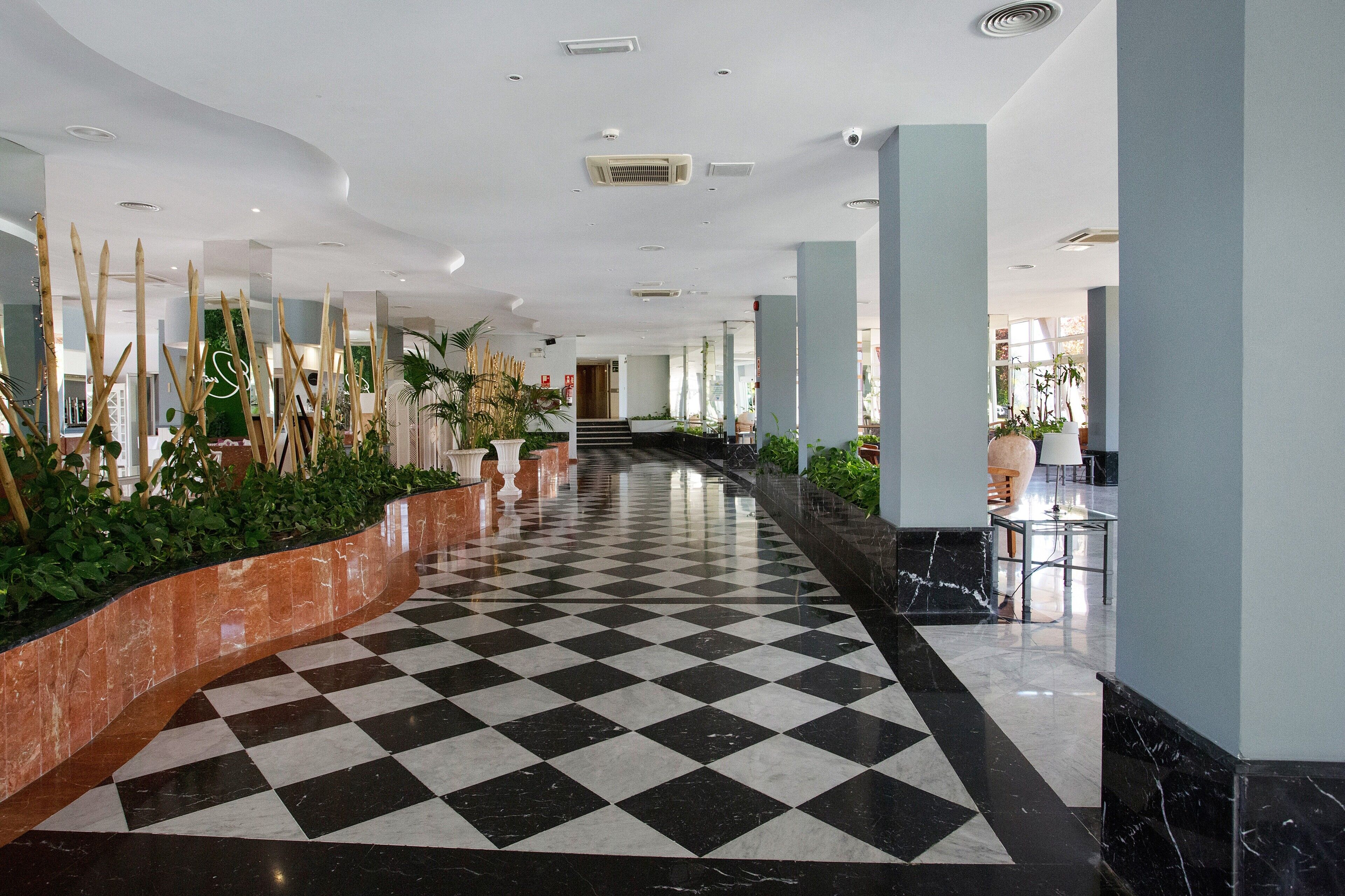 A hotel lobby with a checkered floor, columns, potted plants, and a staircase.