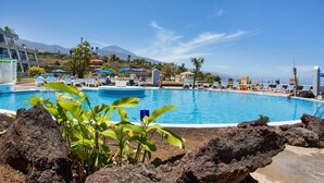A swimming pool with lounge chairs and umbrellas, surrounded by rocks and plants. - Vacation Apartment 'Casa Ana' with Sea and Teide View, Shared Pool and Wi-Fi (SANTA URSULA)