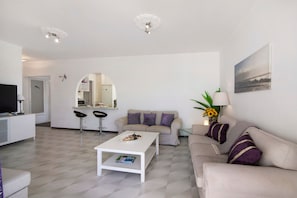 A modern living room with a white coffee table, a beige sofa, and a dining area with a white countertop and bar stools.