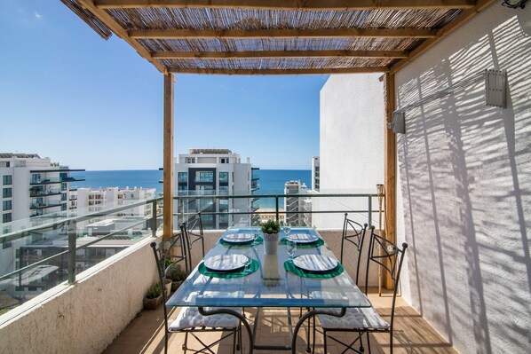A balcony with a glass table set for two, overlooking a cityscape and the sea.