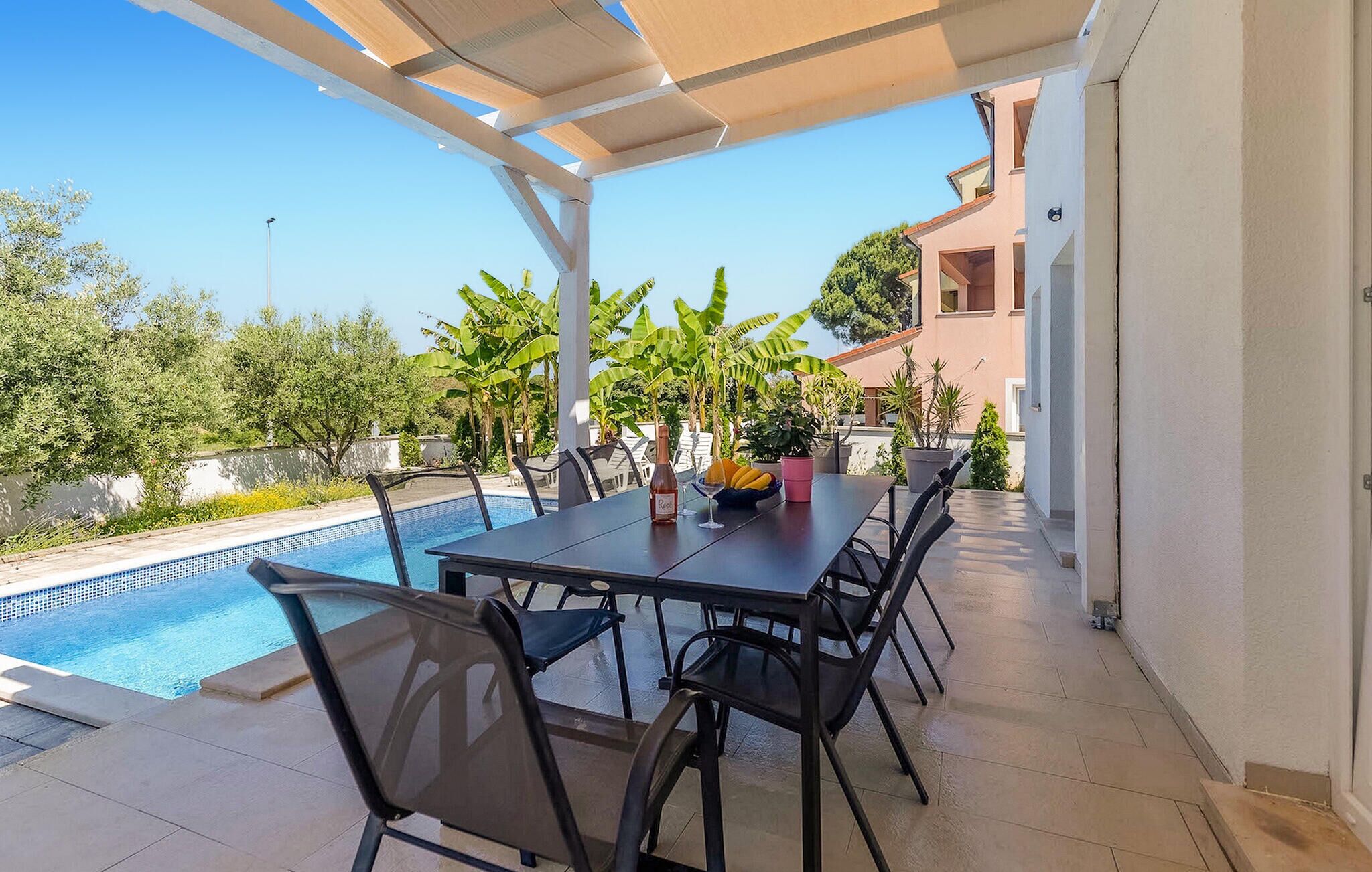 A covered outdoor dining area with a pool, chairs, and a table with a bottle and fruit.