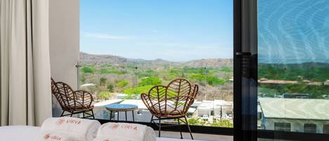 Habitación de hotel con un ventanal grande, dos sillones de mimbre, una mesita y dos toallas con el nombre "Virya".