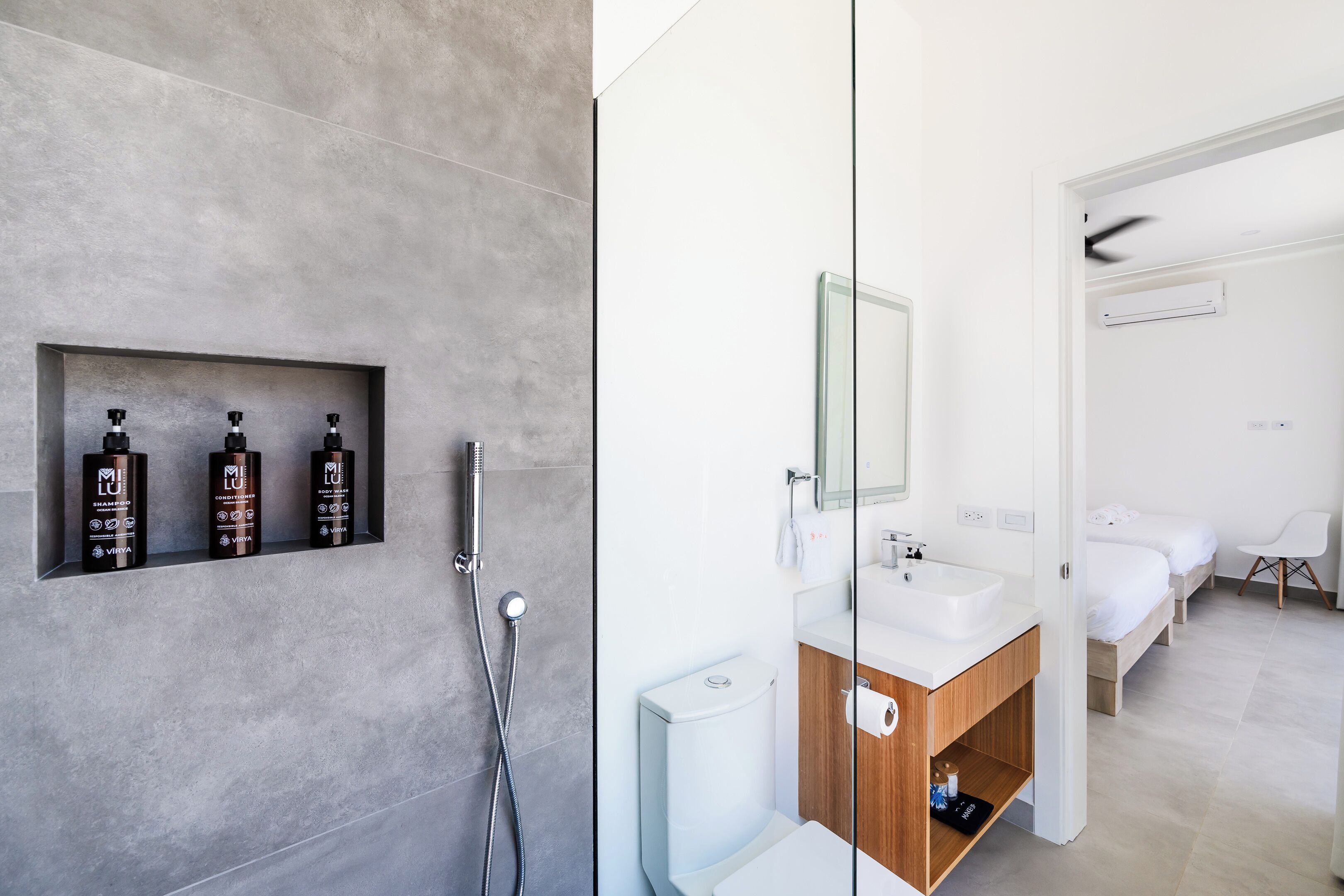 Un baño moderno con zona de ducha con azulejos grises, un lavamanos blanco y un mueble de tocador de madera. Hay tres artículos de aseo en la pared y un espejo sobre el lavamanos.