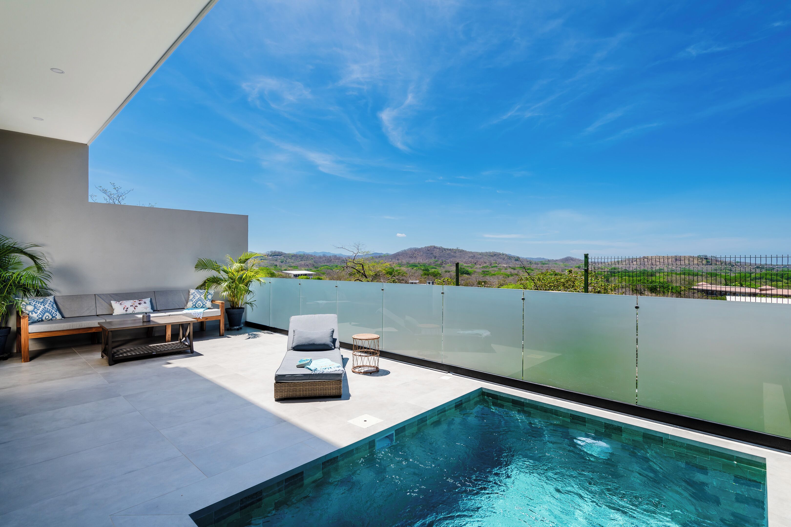 A rooftop pool area with a glass balustrade, a seating area with a sofa and chairs, and a view of the surrounding landscape.
