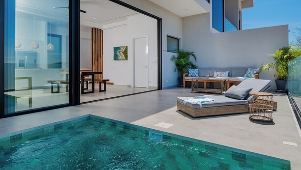 Un área de piscina moderna al aire libre con un porche acristalado, una sala de estar con sofá y mesa, y vistas del edificio contiguo.