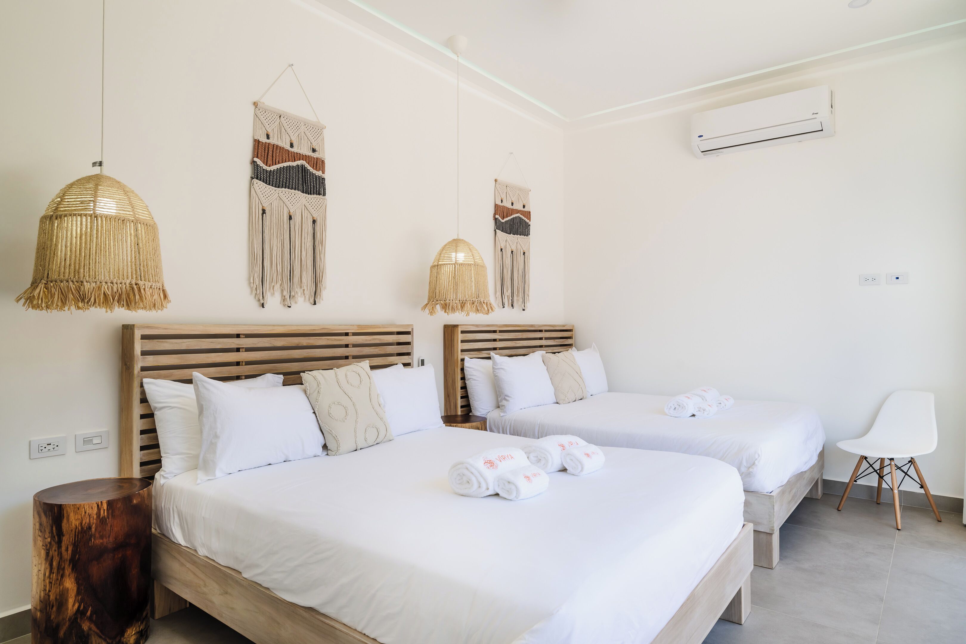 A hotel room with two beds, a wooden nightstand, a woven pendant light, macrame wall hangings, and a white chair.