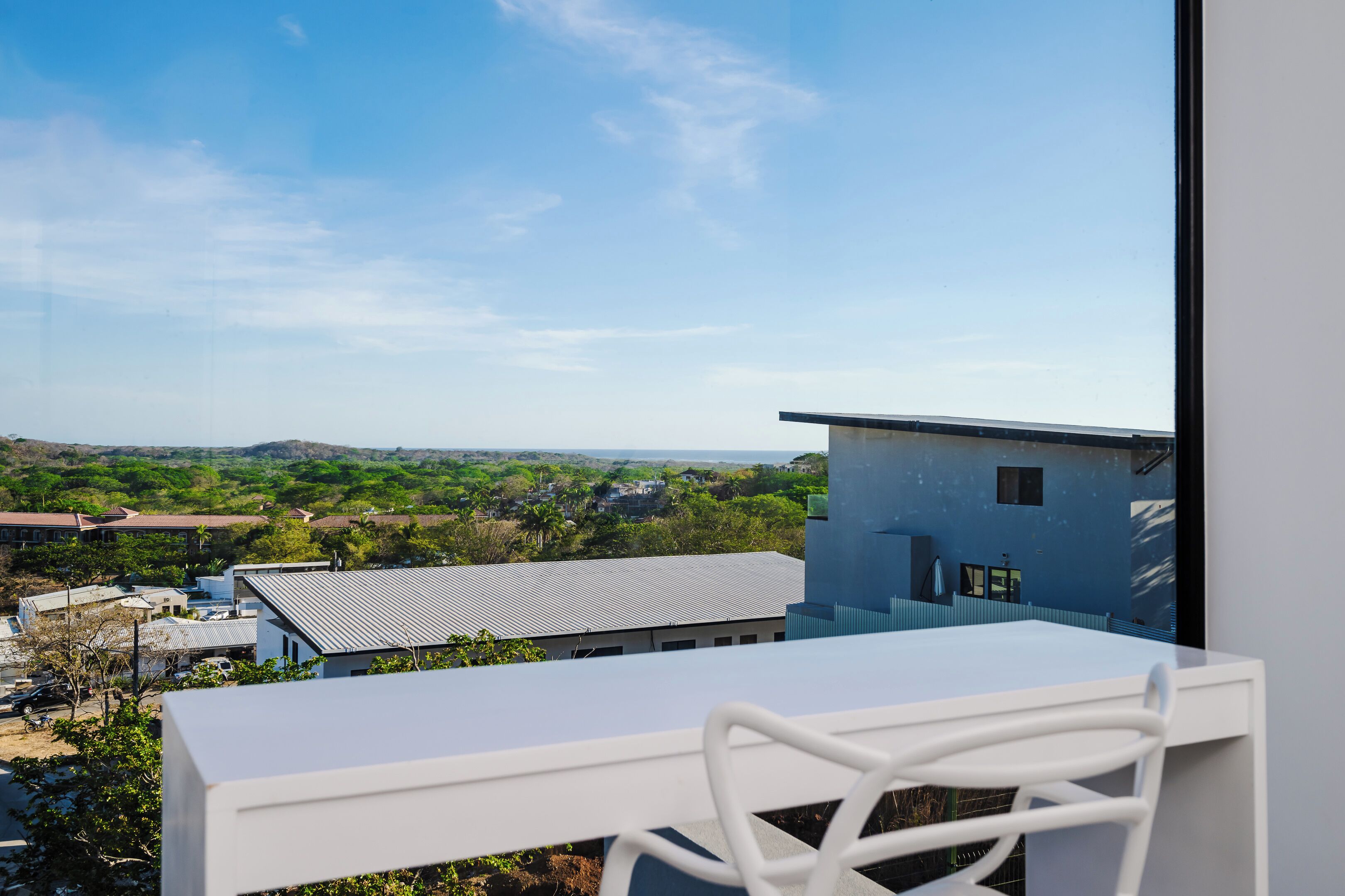 Un balcón con una mesa y una silla blancos, con vista a una zona residencial con casas y vegetación.
