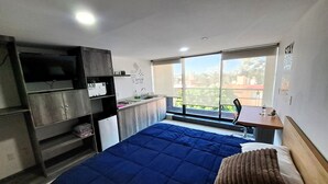 A bedroom with a bed, a desk, a window, and a kitchenette.