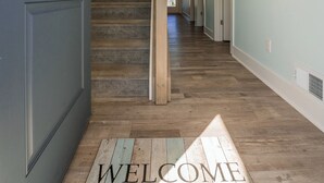 A hallway with a wooden floor, a welcome mat, and a staircase leading to another room. - Anchor's Away | Captain's Cove Waterfront w/ Pool & Golf, Pet Friendly + First-Floor Suite (Greenbackville)