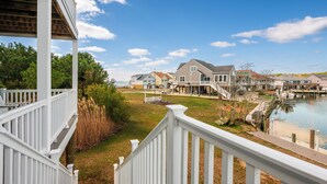 A waterfront property with a wooden deck, a grassy lawn, and a dock extending into the water. - Anchor's Away | Captain's Cove Waterfront w/ Pool & Golf, Pet Friendly + First-Floor Suite (Greenbackville)