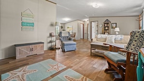 A spacious living room with a wooden floor, a blue armchair, a sofa with beach-themed pillows, a wooden cabinet, and a wall-mounted sign with nautical phrases.