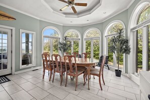 A dining area with a round table and wooden chairs, large arched windows, and potted plants. - Sunset on the Rocks | 4BR Bayview Butler's Bluff Retreat w/ Game Room + Pet Friendly (Cape Charles)