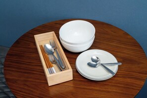 A wooden table with a white bowl, a wooden box containing cutlery, and a stack of white plates.