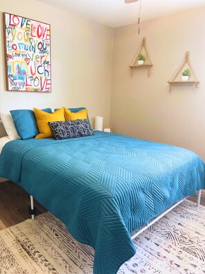 Un dormitorio con colcha color turquesa, un cuadro de arte colorido y dos pequeñas estanterías de madera con plantas.