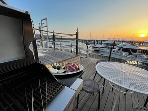 A barbecue grill with various meats and vegetables on a wooden deck overlooking a marina with boats and a bridge in the background.
