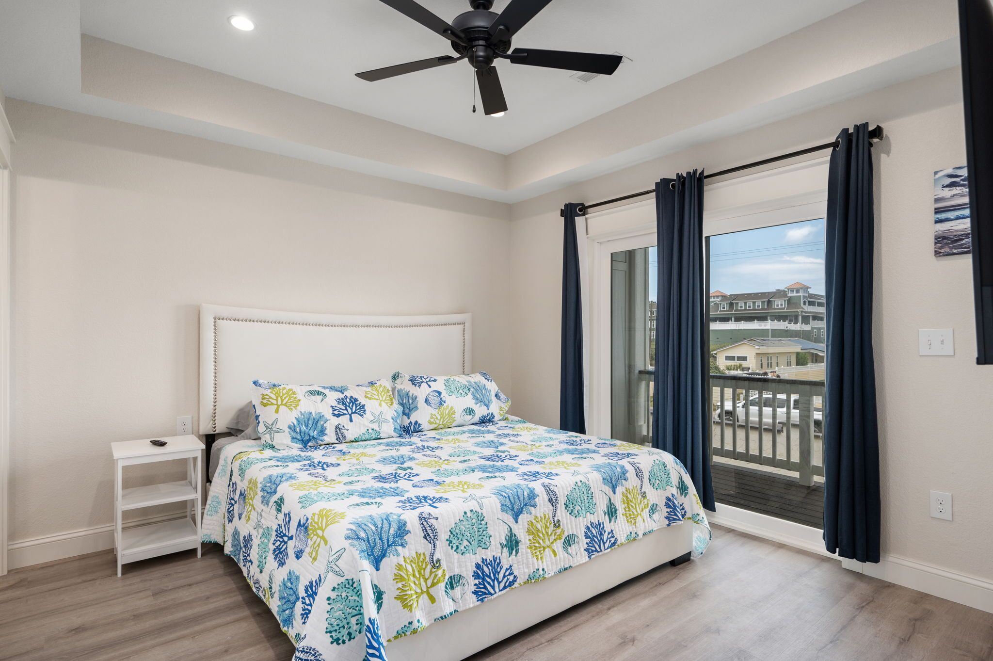 A bedroom with a bed, a nightstand, a ceiling fan, a window with curtains, and a view of buildings outside.