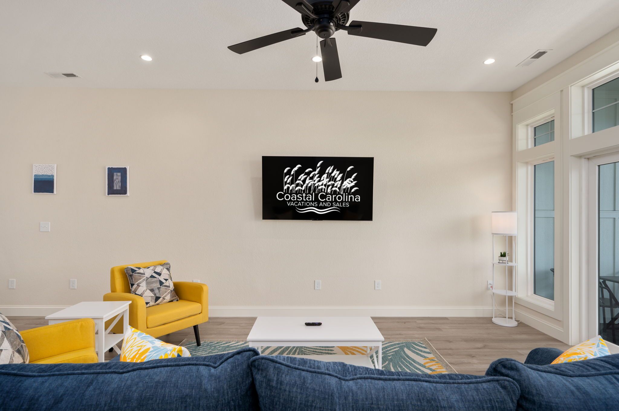 A modern living room with a ceiling fan, a large framed sign, a yellow armchair, a white coffee table, and a blue sofa.