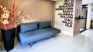 Un moderno vestíbulo de hotel con un sofá gris, una planta decorativa y una pared con saludos en varios idiomas.