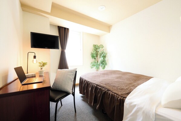 Ein Hotelzimmer mit einem Bett, einem Schreibtisch mit einem Laptop, einem Sessel, einem an der Wand montierten Fernseher, einem Fenster mit Vorhängen und einer Pflanze.