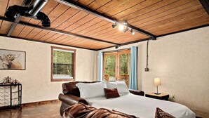 A bedroom with a wooden ceiling, a bed with brown leather headboard, a nightstand with a lamp, and a window with blue curtains.