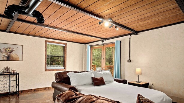 Een slaapkamer met een houten plafond, een bed met een bruin leren hoofdeinde, een nachtkastje met een lamp en een raam met blauwe gordijnen.