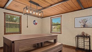Een kamer met een houten plafond, een grote tafel met banken, een wandklok en een raam met jaloezieën.