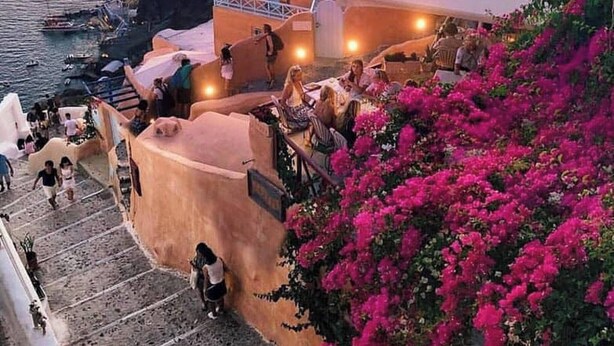 Un edificio costero con una escalera que conduce a una terraza con vista al mar, rodeado de flores de buganvilla rosadas.