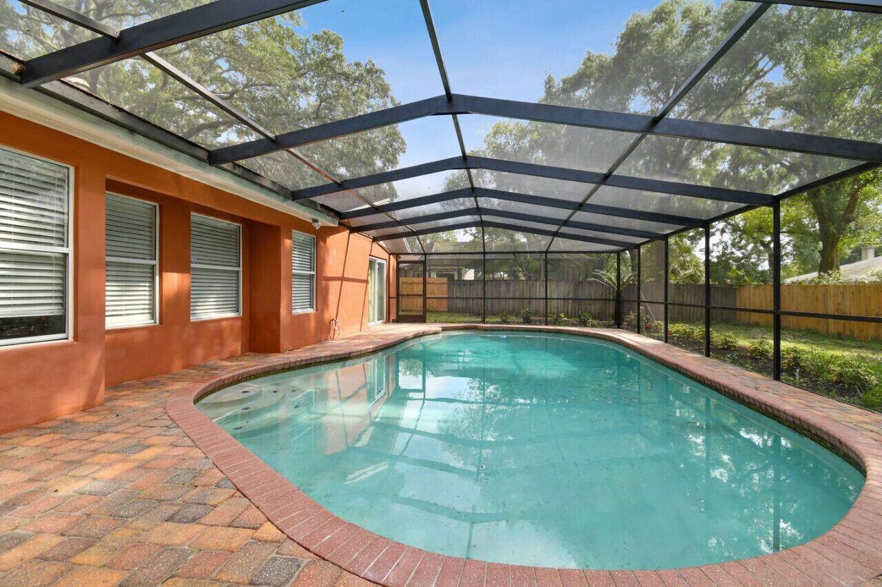 A covered swimming pool with a glass ceiling, surrounded by a brick patio and a wooden fence.