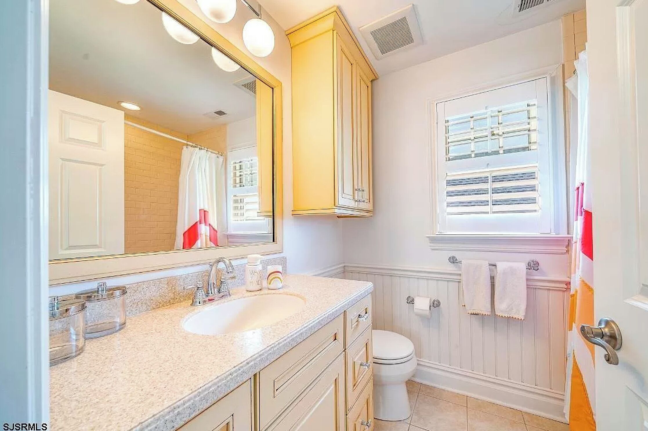 A bathroom with a white sink, vanity, and toilet. There is a window with blinds and a towel rack with towels.
