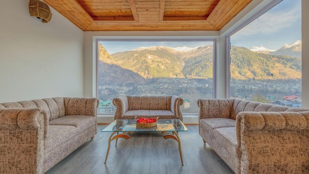 A room with a wooden ceiling, two patterned sofas, a glass coffee table, and a large window with a mountain view. - Hyacinth Hotel (Manali)