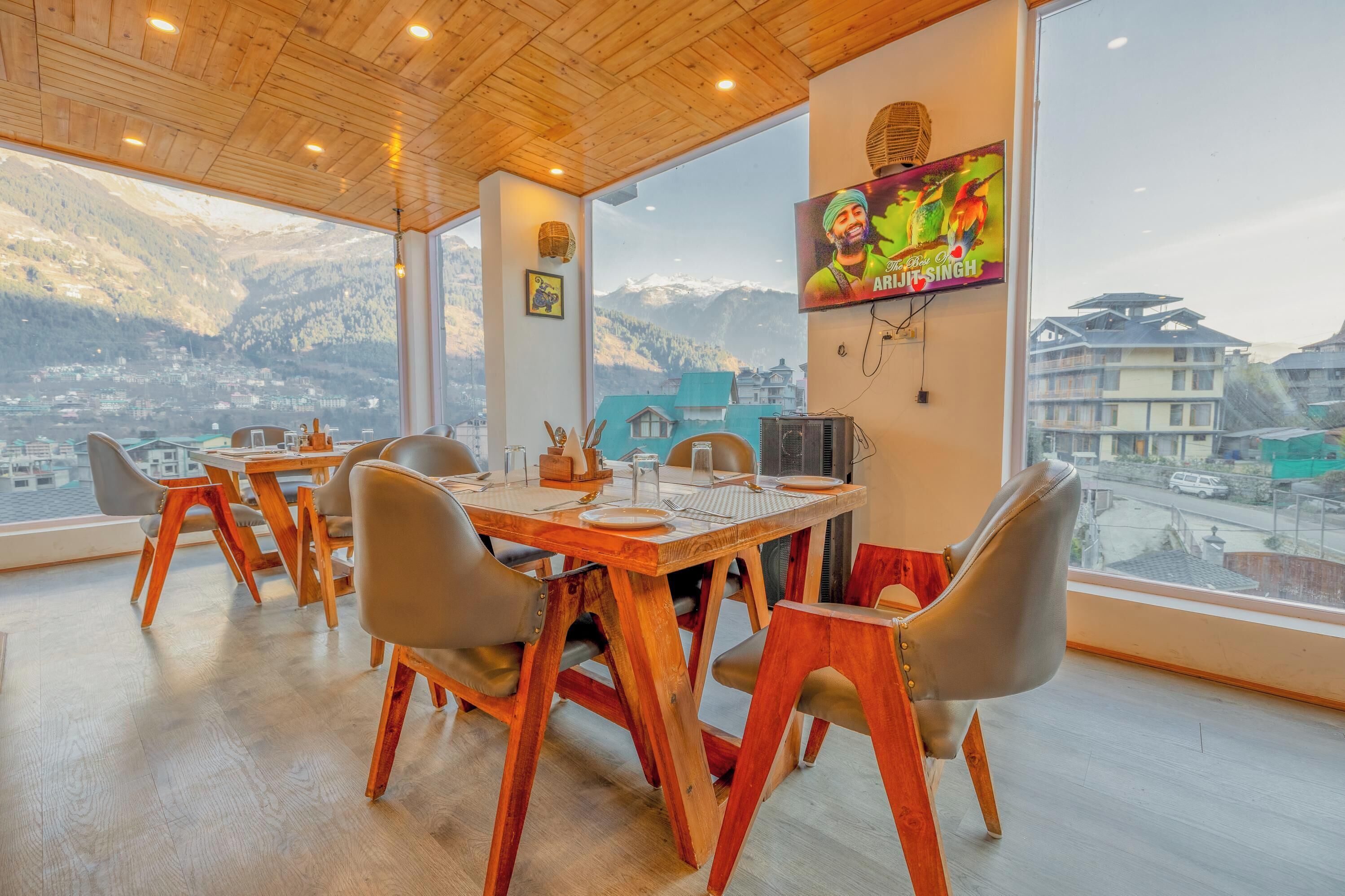 A dining area with a wooden table and chairs, a TV mounted on the wall, and a view of mountains through large windows.
