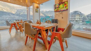 A dining area with a wooden table and chairs, a TV mounted on the wall, and a view of mountains through large windows. - Hyacinth Hotel (Manali)