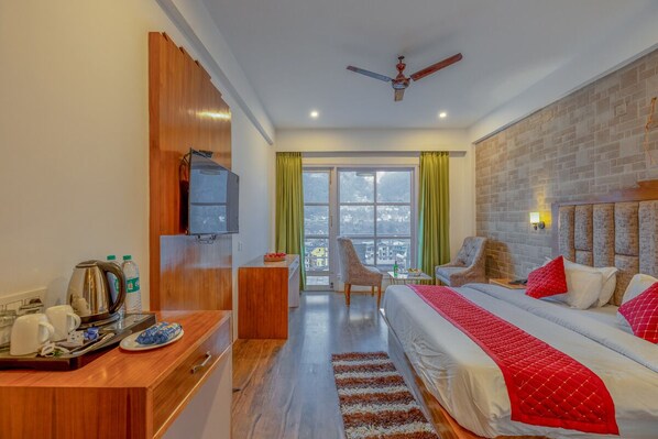 A hotel room with a bed, a desk with a kettle and cups, a chair, and a view of the city through large windows. - Hyacinth Hotel (Manali)