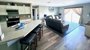 A modern kitchen with a central island, a dining area with bar stools, a living room with a sofa and a coffee table, and a large window with curtains.