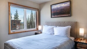A bedroom with a large bed, two bedside tables, a window with a view of trees and a building, and a framed picture on the wall.