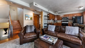A spacious living room with a brown leather sofa set, a coffee table with magazines, and a kitchen in the background.