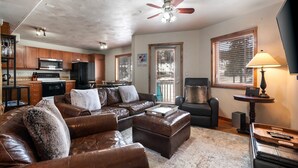 A spacious living room with a brown leather sofa, a coffee table, and a TV stand. There is a kitchen area with a microwave, stove, and refrigerator.
