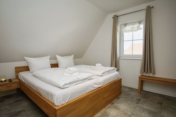 A neatly made bed in a room with a sloped ceiling, a bedside table with a lamp, and a window with curtains.