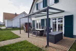 Un patio couvert avec une table et des chaises, un barbecue et un chemin qui traverse le jardin.