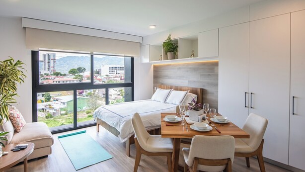 Ett modernt hotellrum med en stor säng, ett matbord och stolar, utsikt över staden genom stora fönster och en liten balkong.