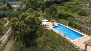 En swimmingpool omgivet af frodigt grønt og stenvægge, med en sti der fører hen til en bygning i baggrunden.