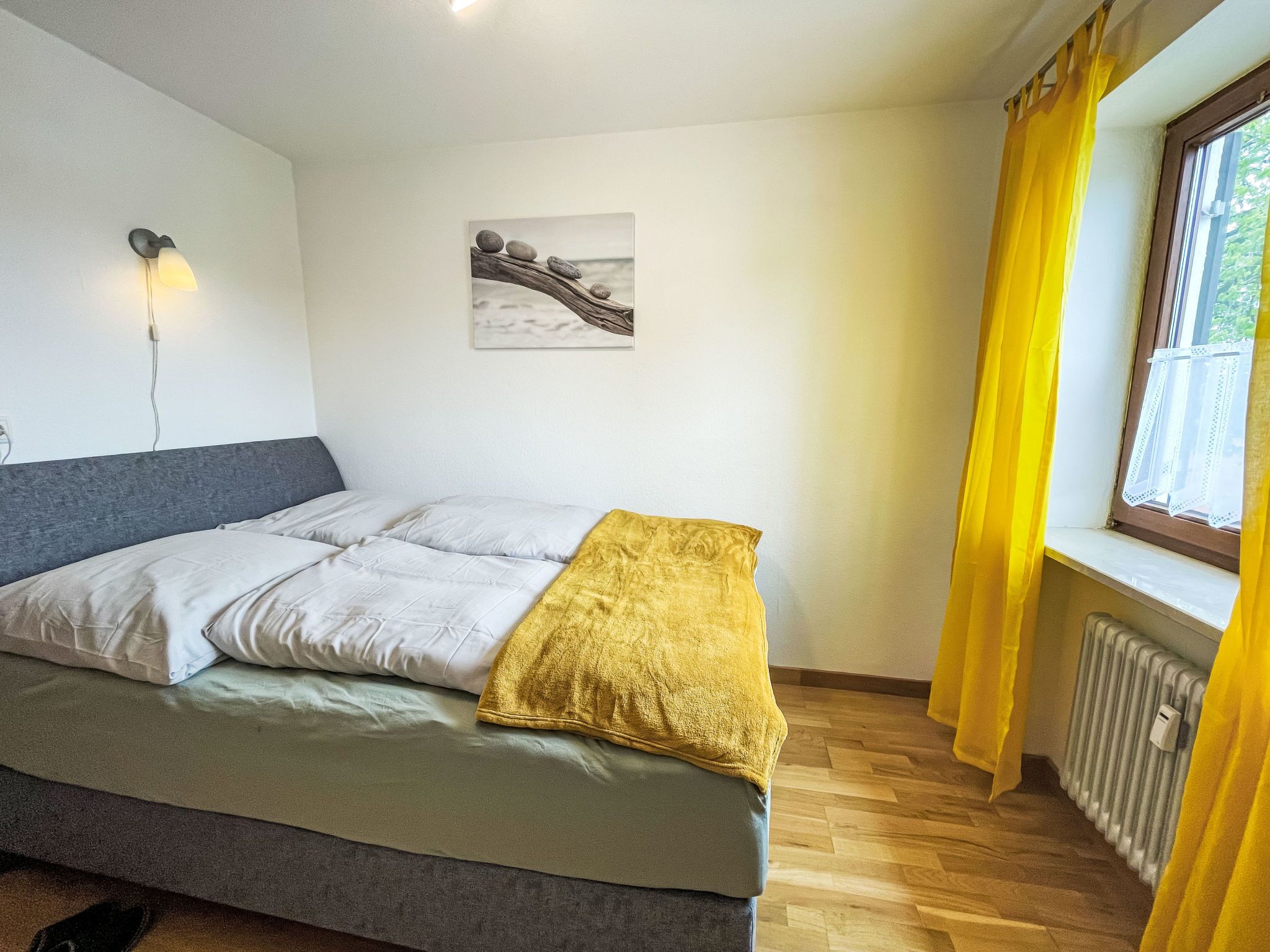 Een slaapkamer met een bed, een wandlamp, een schilderij en een raam met gele gordijnen.