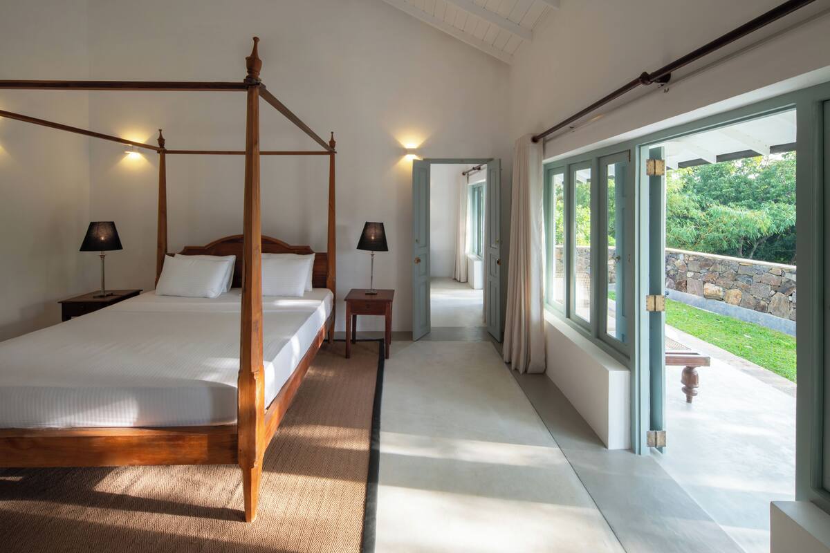 A spacious bedroom with a four-poster bed, a nightstand, a bedside table, and a view of a garden through large windows.