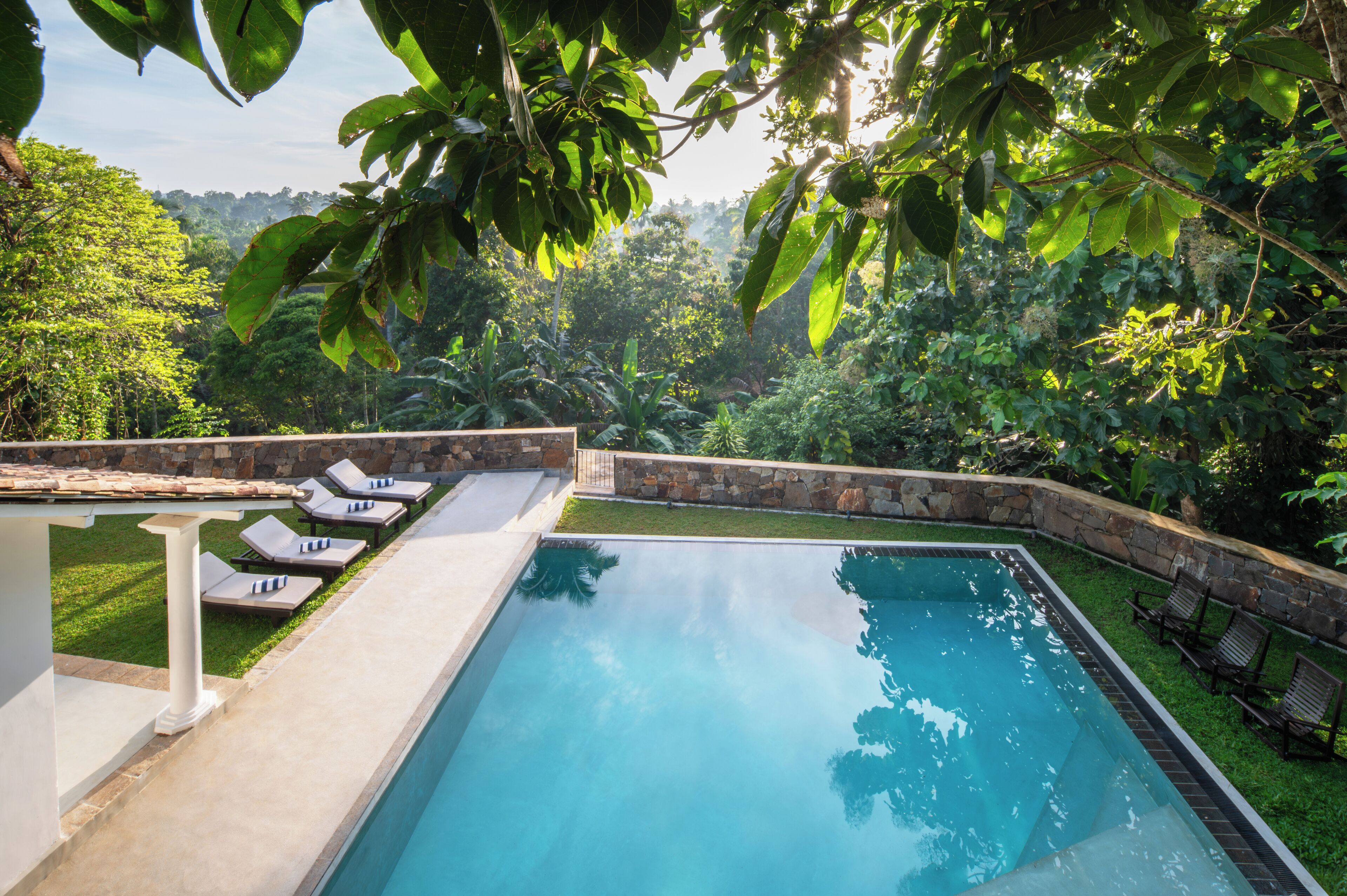 A rectangular swimming pool with a stone border, surrounded by a stone wall and a white pergola.