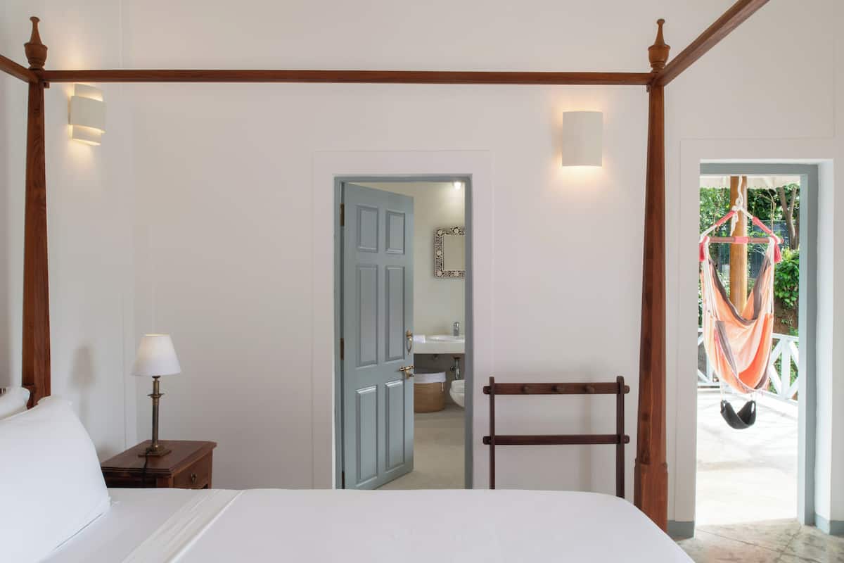 A bedroom with a four-poster bed, a nightstand with a lamp, a wooden chair, and a view of a bathroom and outdoor hammock.