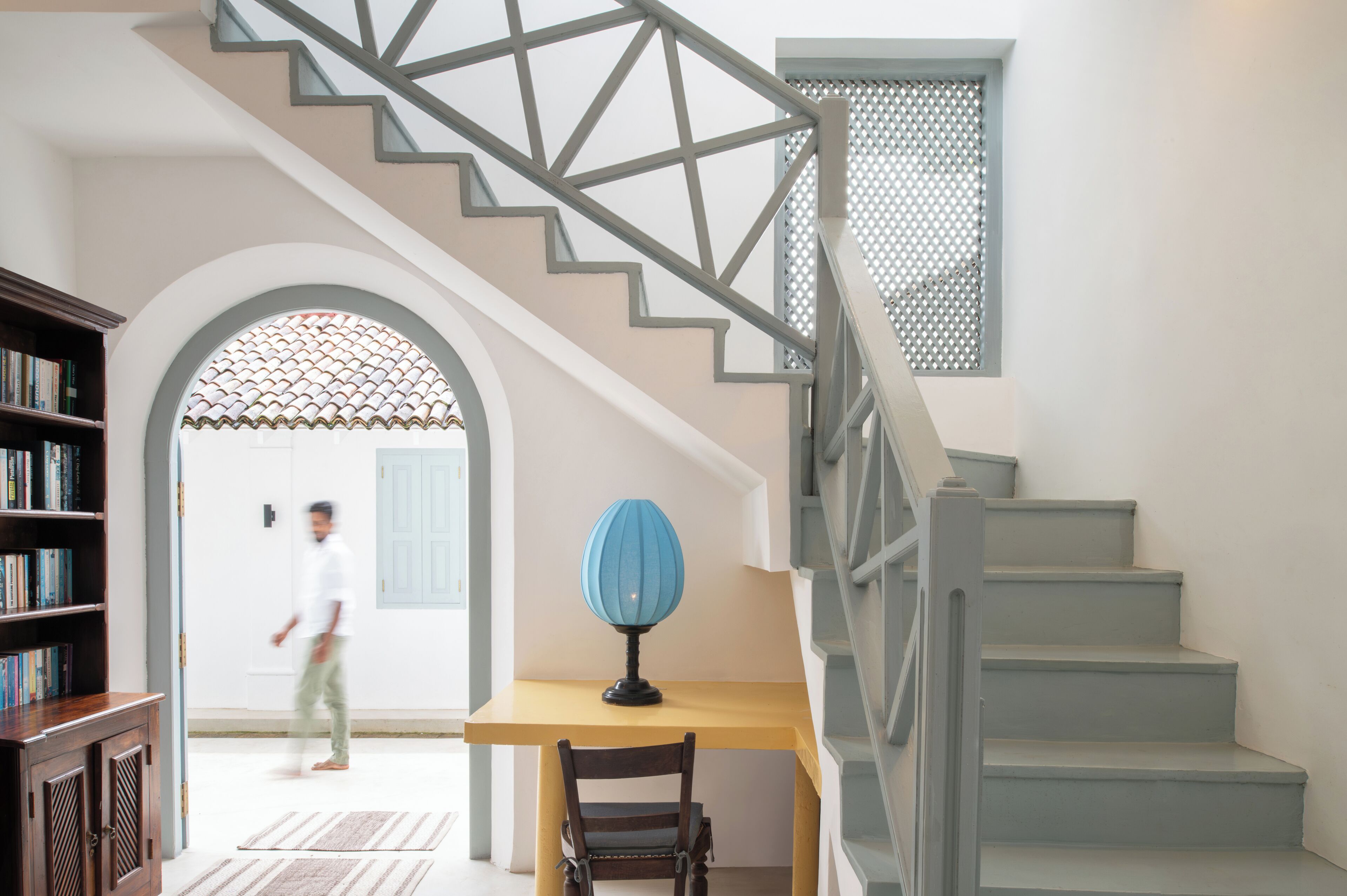 A staircase with a geometric railing design, a wooden table with a blue lamp, and a bookshelf with books.