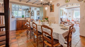 Un coin repas avec une grande table, des chaises en bois et un lustre. On aperçoit une cuisine au fond.