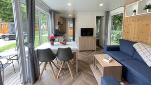 A modern living room with a sofa, a coffee table, and a dining area with chairs and a table. There is a TV on a wooden cabinet, and a kitchen area is visible in the background.