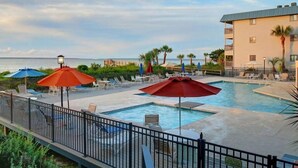 A pool area with lounge chairs and umbrellas, a building with balconies, and a view of the ocean. - Cozy Beach and Racquet Condo | Heated Pool Access (Tybee Island)